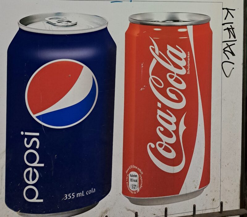 Lata de Pepsi azul a la izquierda junto a una lata de Coca-Cola roja a la derecha, simbolizando la clásica guerra de las colas y la competencia en el mercado de refrescos